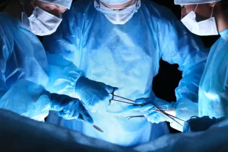 Three surgeons in blue scrubs perform fistula surgery on a patient in a sterile operating room.
