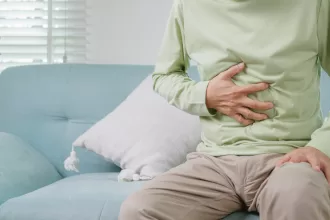 A man sits on a couch, grimacing in pain, indicating discomfort likely due to a hernia condition requiring surgery.