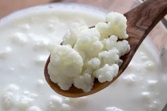 A glass of creamy white kefir on a wooden table, with whole berries, grains, and a heap of kefir grains next to it, illustrating the benefits of the kefir to the health.